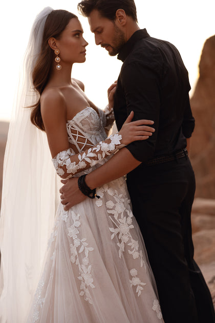 The Nabila wedding dress with a lace corset bodice and off-shoulder sleeves highlights feminine curves with intricate floral embroidery.
