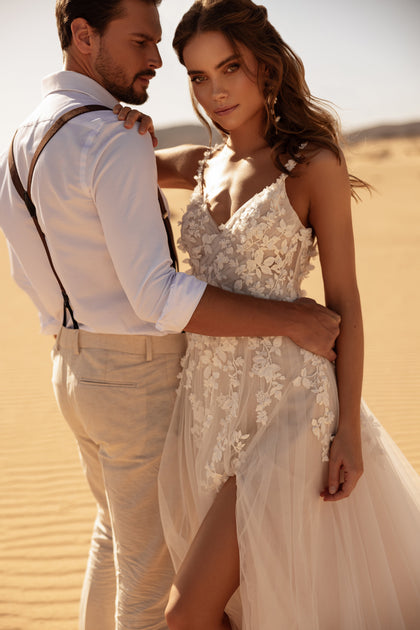 The Aviva wedding dress features a sheer lace bodice with 3D floral details, highlighting the bride’s figure in the desert light.
