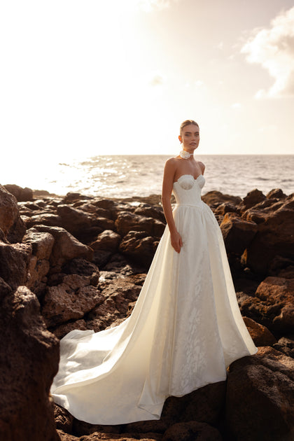 The Cholly wedding dress with a structured bustier bodice and long train, captured at sunset by the sea, embodies timeless bridal elegance.