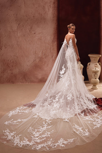 The Aspasia wedding gown with a dramatic lace train and floral-embroidered cape creates an unforgettable bridal silhouette.