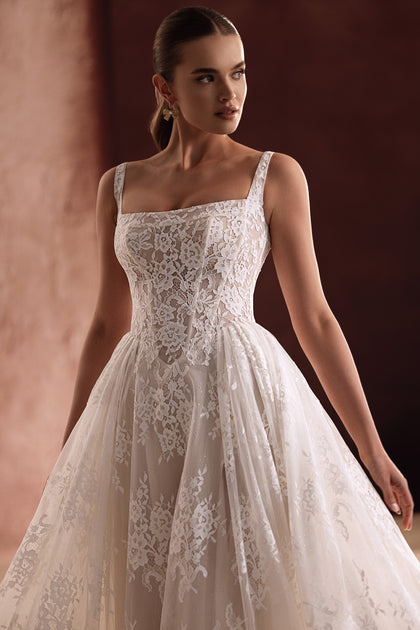 The Becco wedding gown with a square neckline and intricate lace detailing enhances the bride’s silhouette with refined elegance.