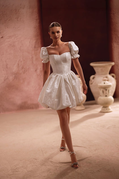 The Daphne short wedding dress showcases a lace-up corset back with puff sleeves, blending romance with modern simplicity.
