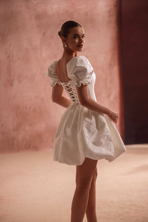 The Daphne wedding mini dress with lace-up corset back and puff sleeves highlights the waist, offering a flirty princess look.