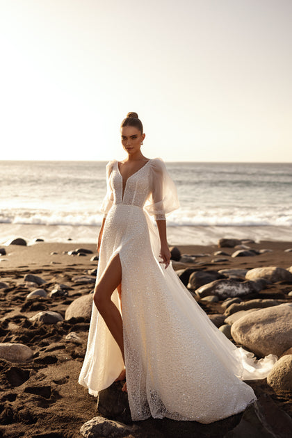 The Davina wedding dress features a plunging V-neckline and shimmering fabric, glowing beautifully in the golden beach sunset.