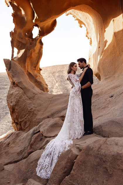 The Dune wedding dress features a sheer lace bodice and bell sleeves with intricate floral embroidery for a romantic boho look.