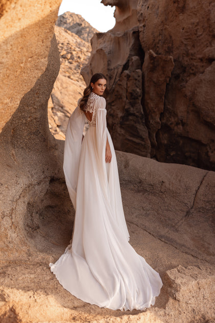 The Etra wedding gown features a deep V-neckline, dramatic slit, and chiffon cape sleeves for a boho-chic bridal look.