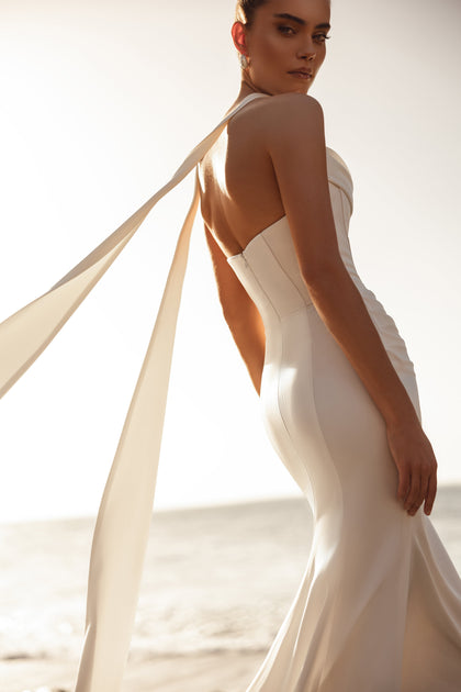 The Luna wedding gown’s long train and flowing ribbons create a striking modern bridal look by the ocean.