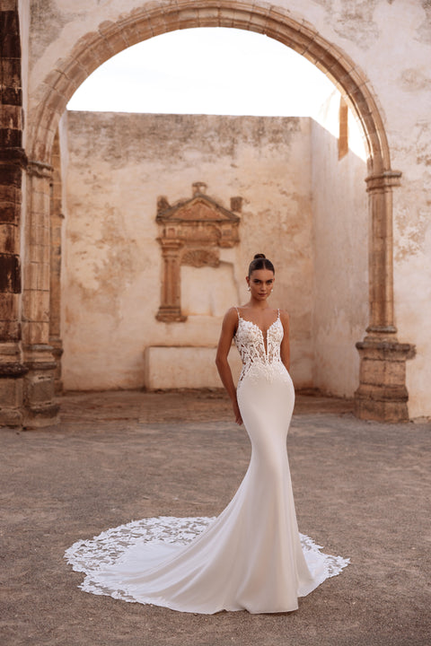 The Munira wedding dress features a deep V-neck lace bodice with intricate embroidery and a figure-hugging mermaid silhouette.