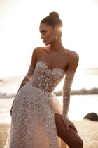 The Reem wedding dress with a corset bodice and high slit dazzles with 3D floral embroidery and shimmering details in the golden sunset light.