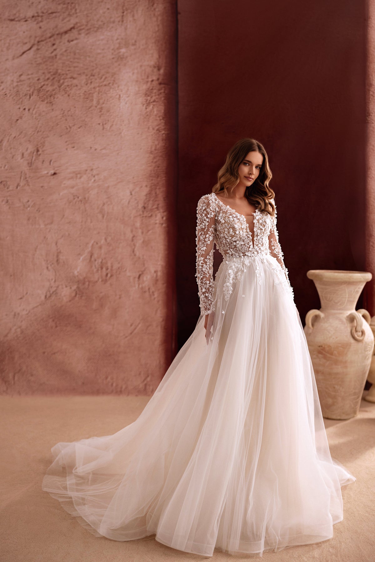 The Shanin wedding dress with long lace sleeves, deep neckline, and flowing tulle skirt exudes effortless romance and modern elegance.